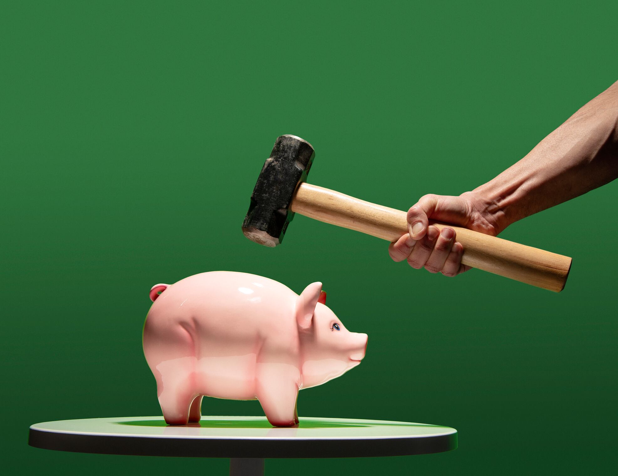 Man's hand about to hit ceramic coin piggy bank with sledge hammer, bank on white table, green back ground.