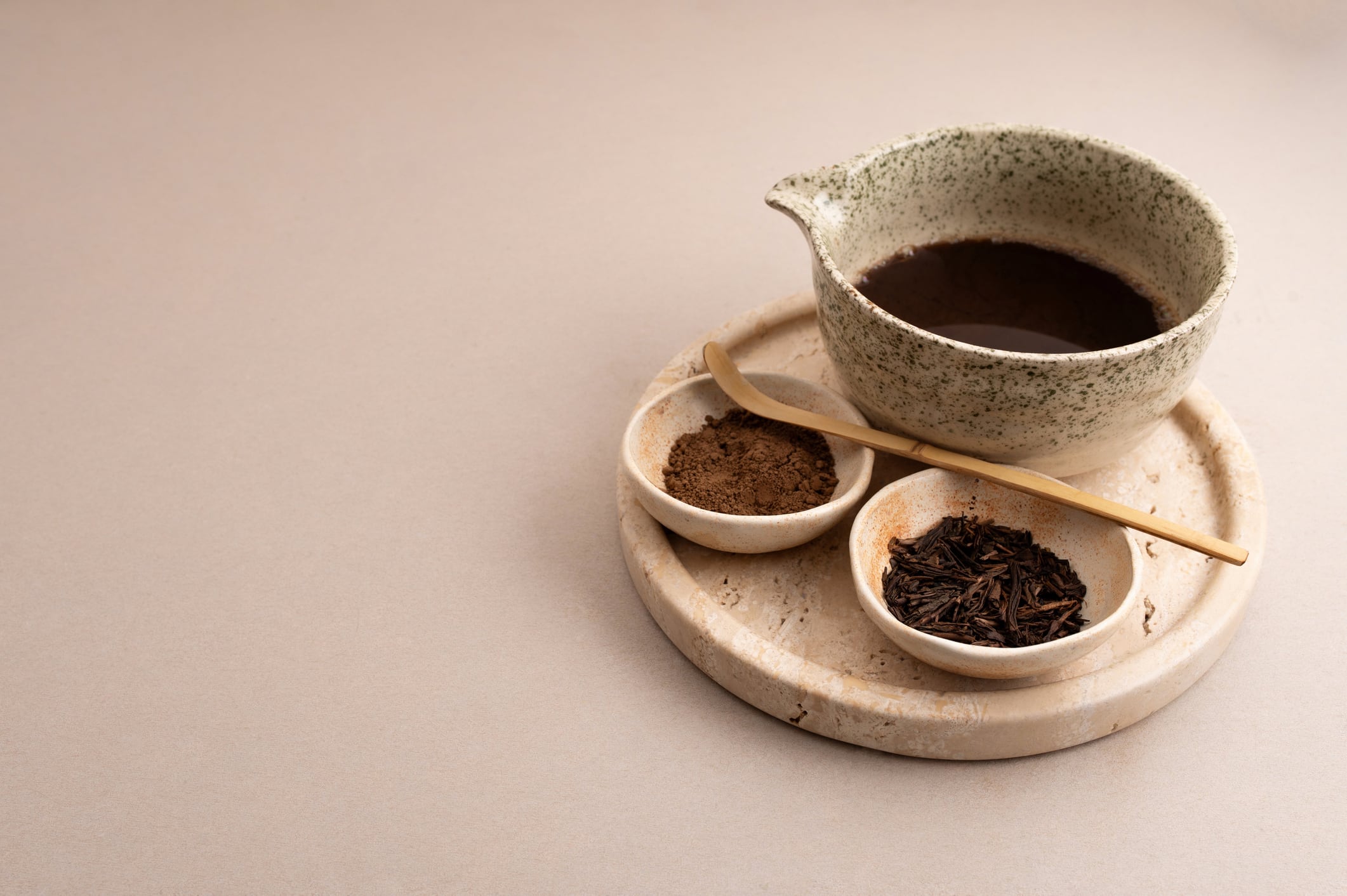 Minimalist horizontal composition of hojicha latte in a sculptural glass with roasted green tea leaves and hojicha powder on a stone tray.