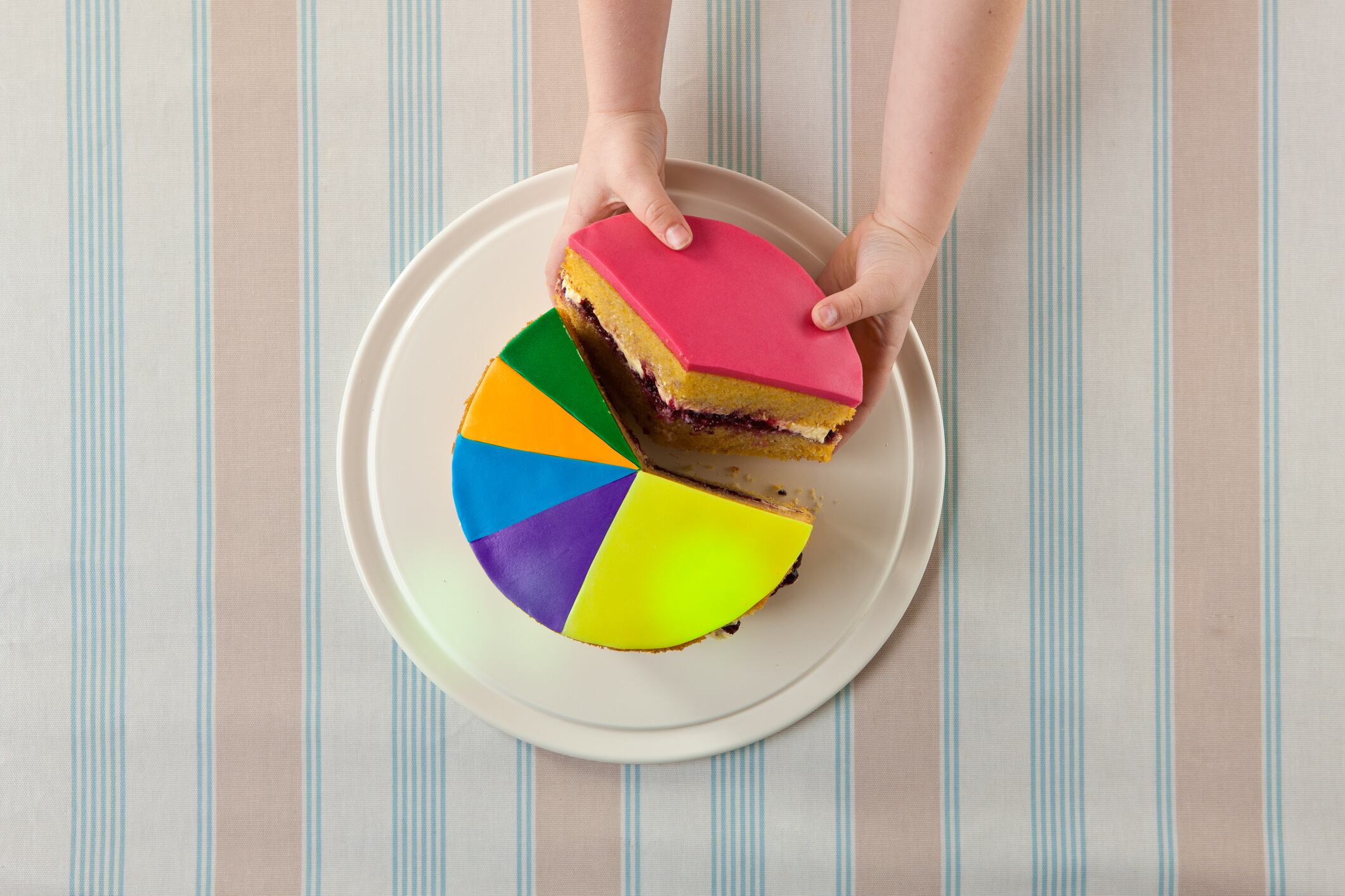 Aerial view of a 6 year old child's hands removing the largest slice of a sponge cake designed as a pie chart.