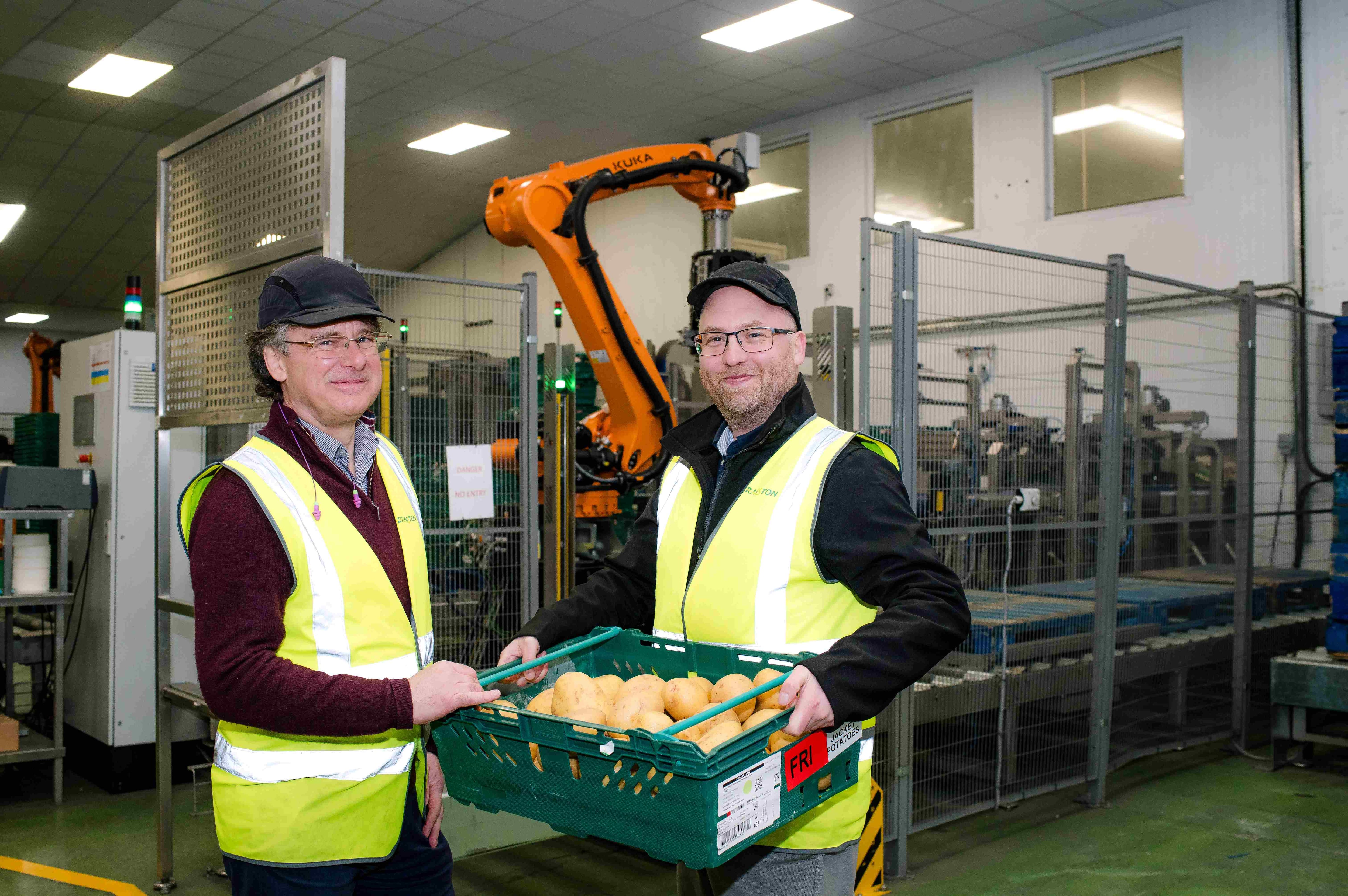 Two men in high vis carrying potatoes in a crate in a factory.
