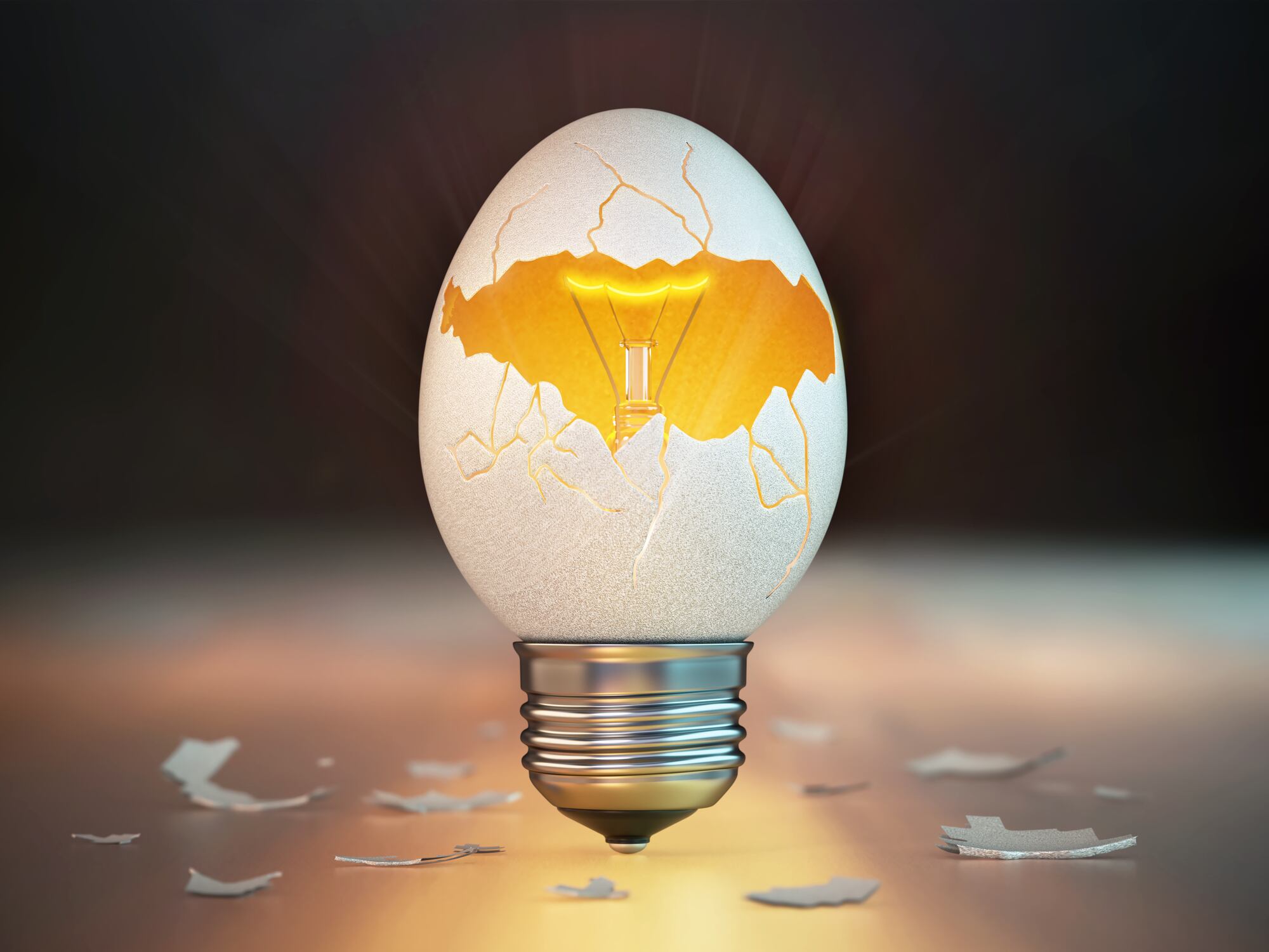 A broken egg shows a light bulb inside. The light shines brightly from the center. Pieces of the eggshell scatter around on the surface nearby. The setting is simple and dark.