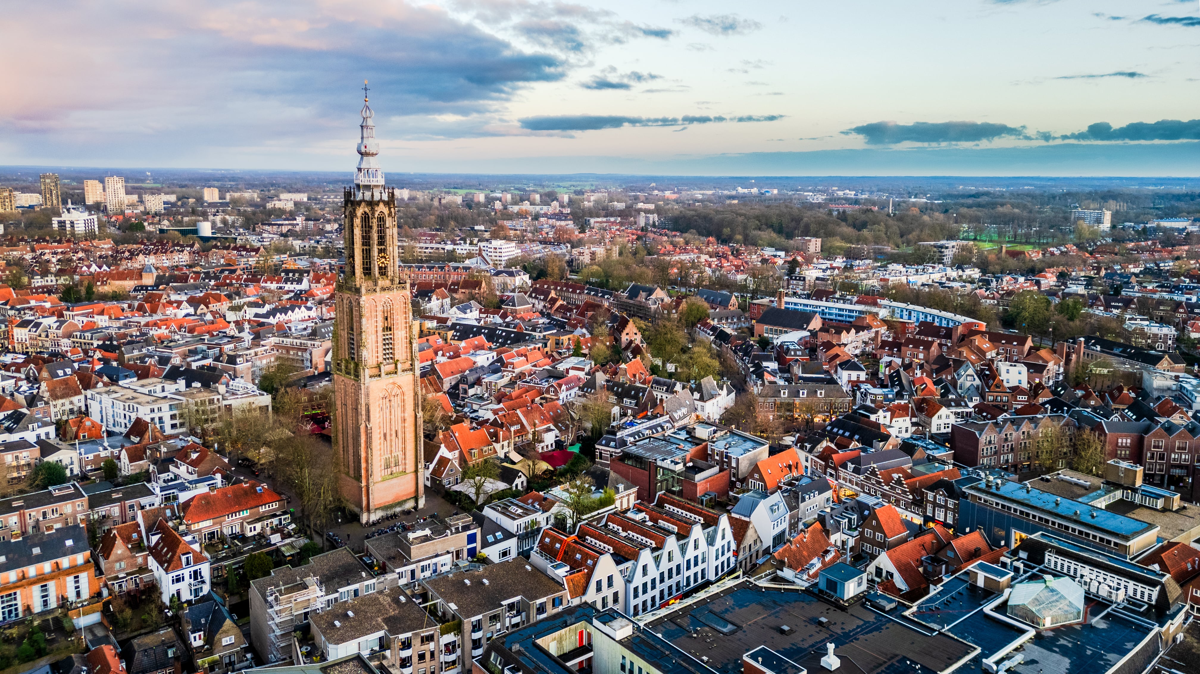 Amersfoort, The Netherlands. Captured at sunset, this aerial view showcases the city’s historic architecture, including the iconic Onze Lieve Vrouwetoren in the center. Surrounding the tower are modern office buildings, residential areas, and railway tracks leading to the station in the foreground. The warm colors of the setting sun highlight the charm of this Dutch cityscape