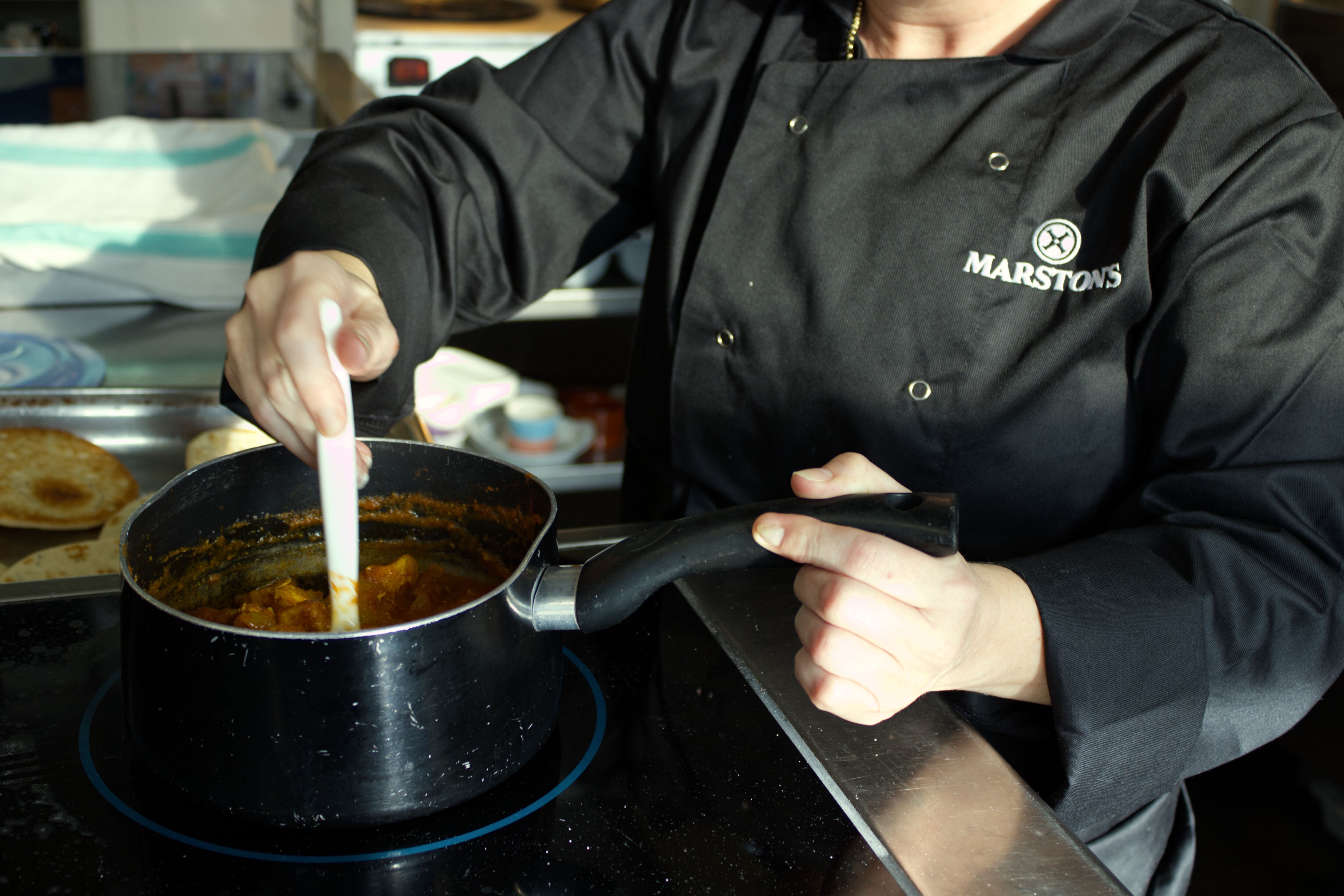 A woman stirring a pot with food and a Marston's branded overall.