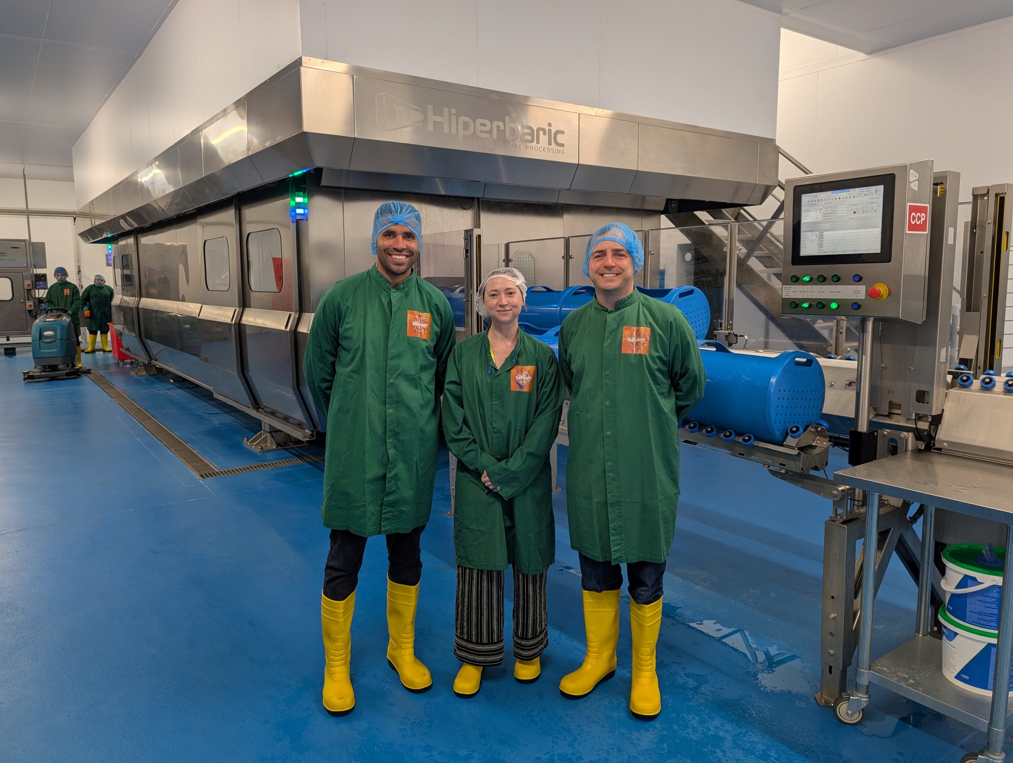 Thomas Robson-Kanu, founder and CEO of The Turmeric Co. ; Bethan Grylls, editor of Food Manufacture; and Ross O'Hara, operations director at The Turmeric Co., dressed in PPE and standing in front of a large machine.