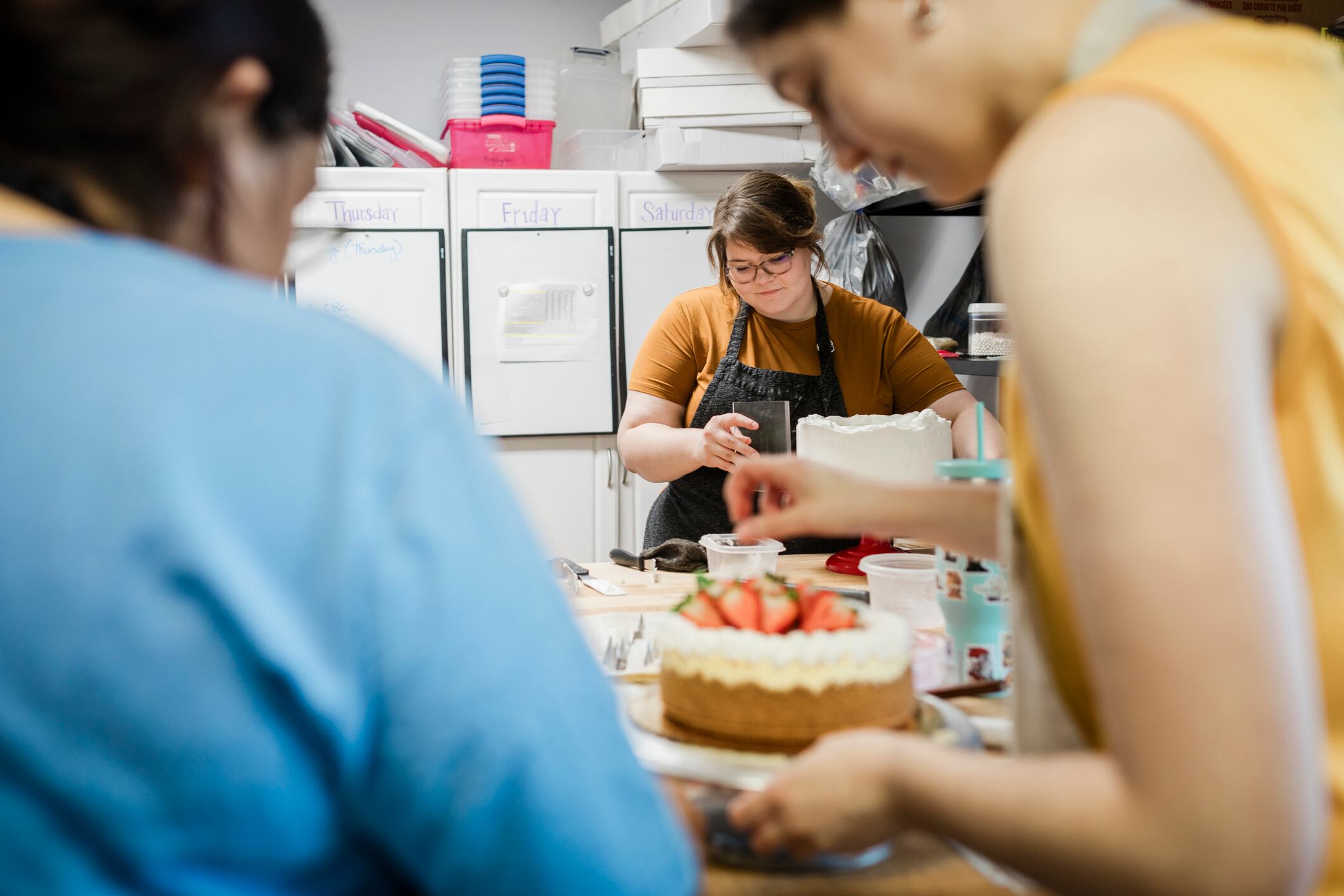 This image captures a team of bakers collaborating in a professional kitchen as they decorate cakes. The focus is on the detailed work of placing fresh strawberries on a cheesecake, with other team members visible in the background, highlighting the cooperative and skilled environment of the bakery.