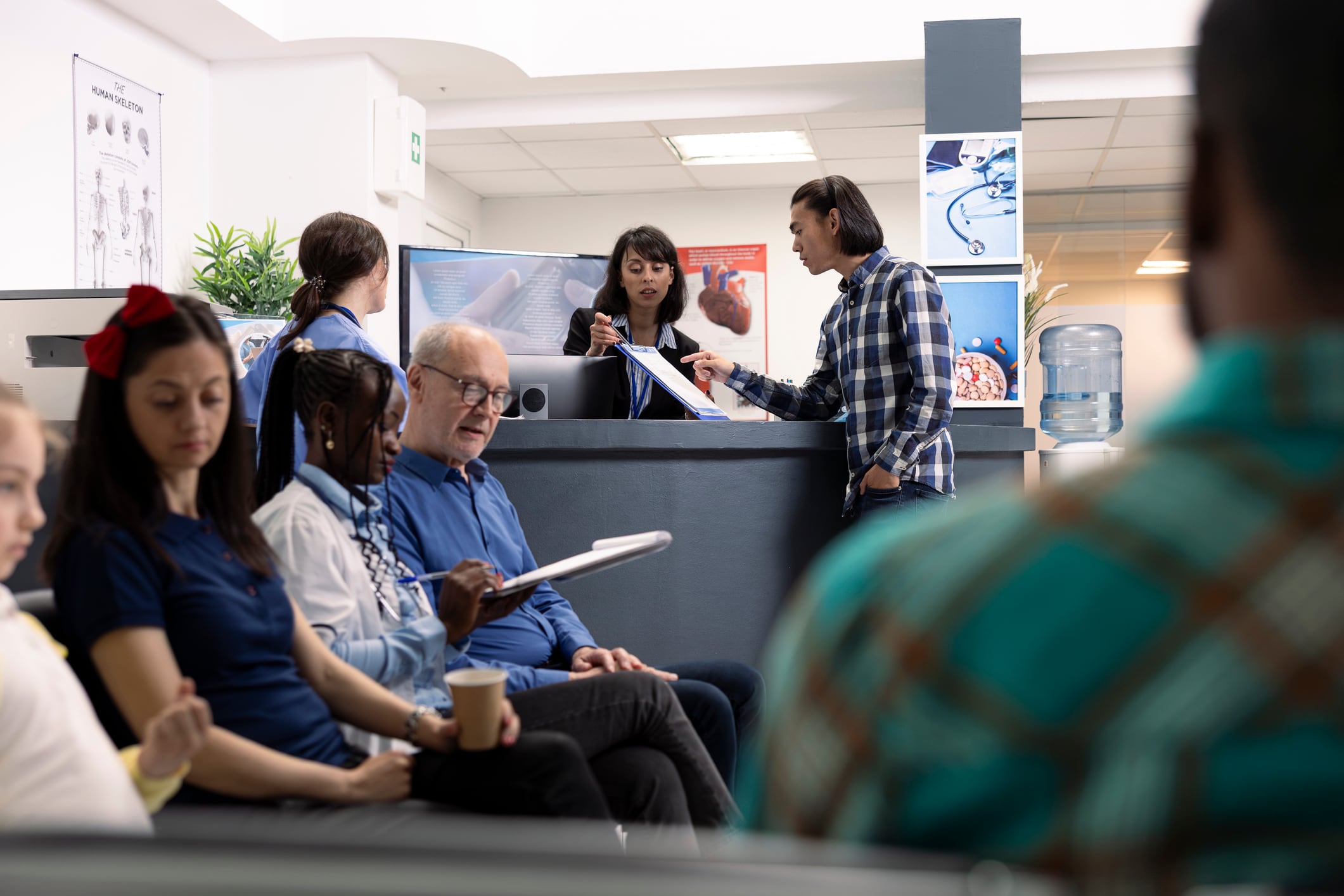 Asian male patient completes medical registration as hospital receptionist explains clipboard healthcare forms in clinic lobby. Young man preparing for doctor appointment in emergency waiting room.