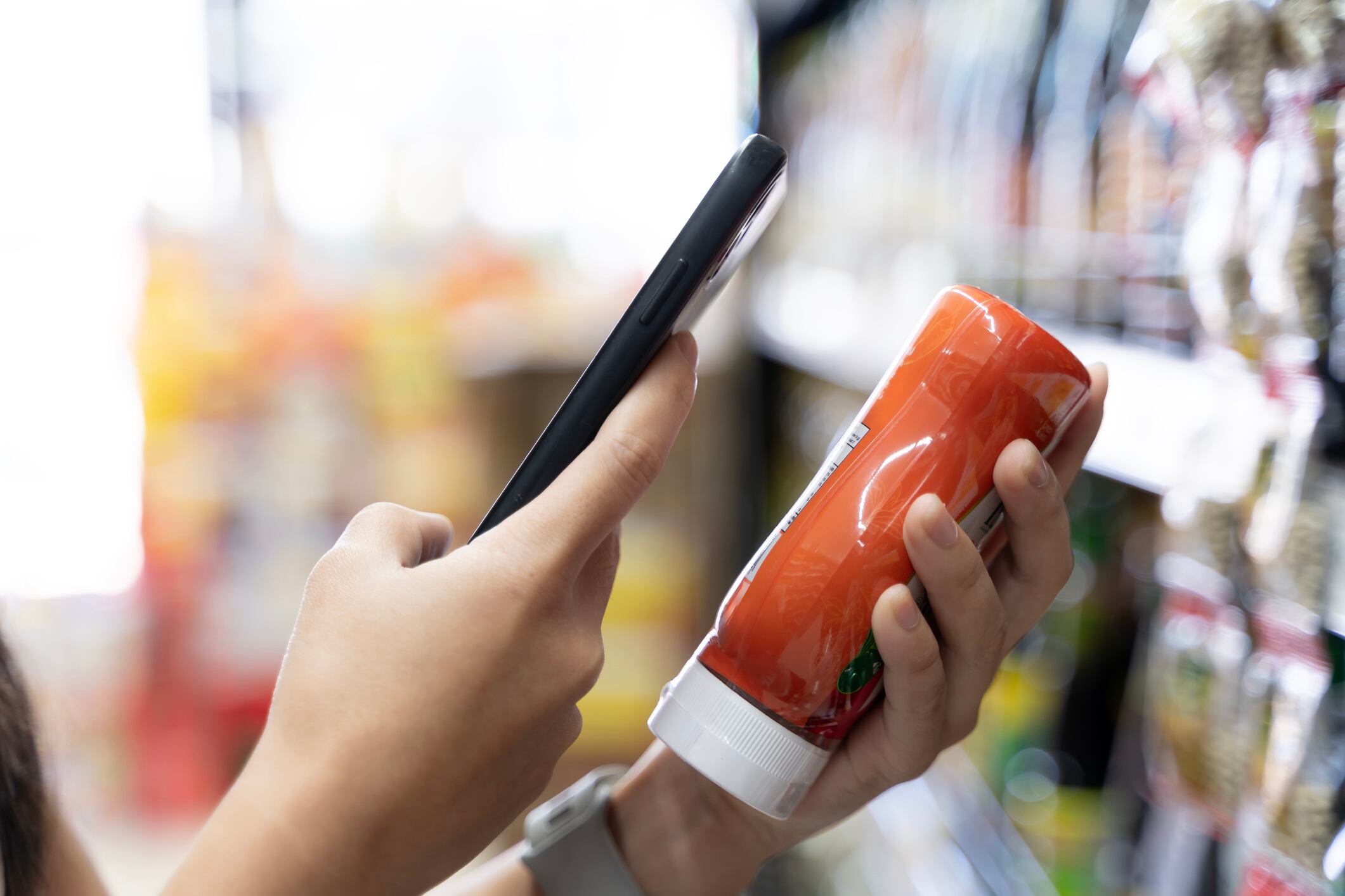 Close-up of a shopper using a smartphone to scan and check product details in a supermarket aisle, highlighting modern retail behavior, digital shopping habits, and consumer decision making.
