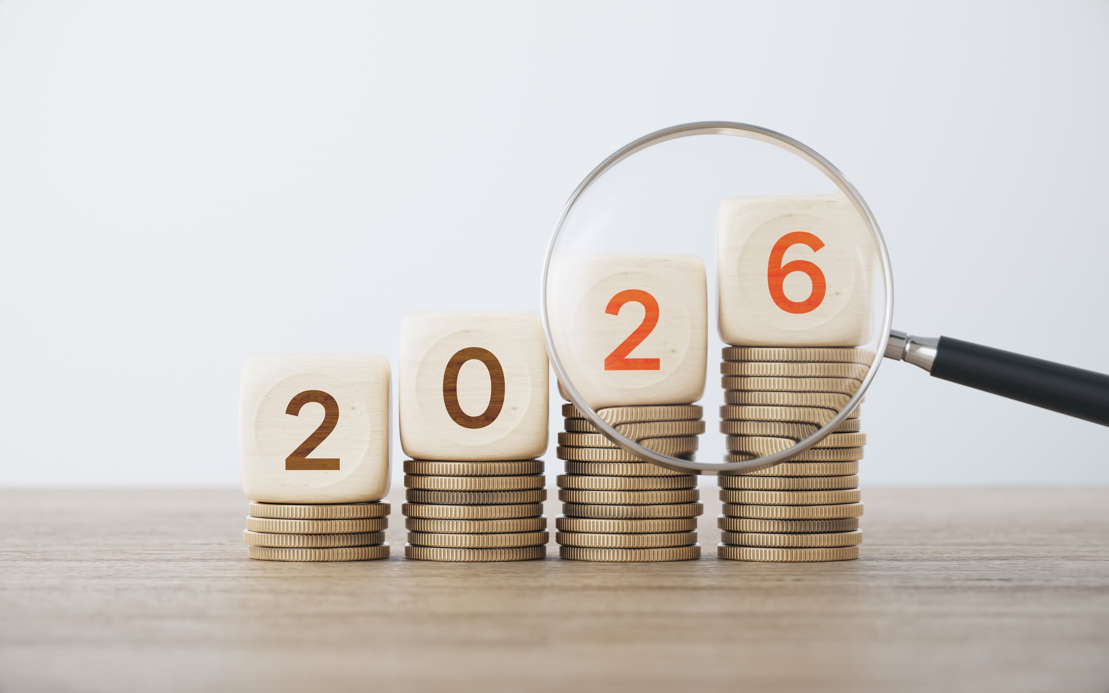 2026 Written Wooden Blocks with Coin Stacks and Magnifying Glass on Defocused Wood Background.