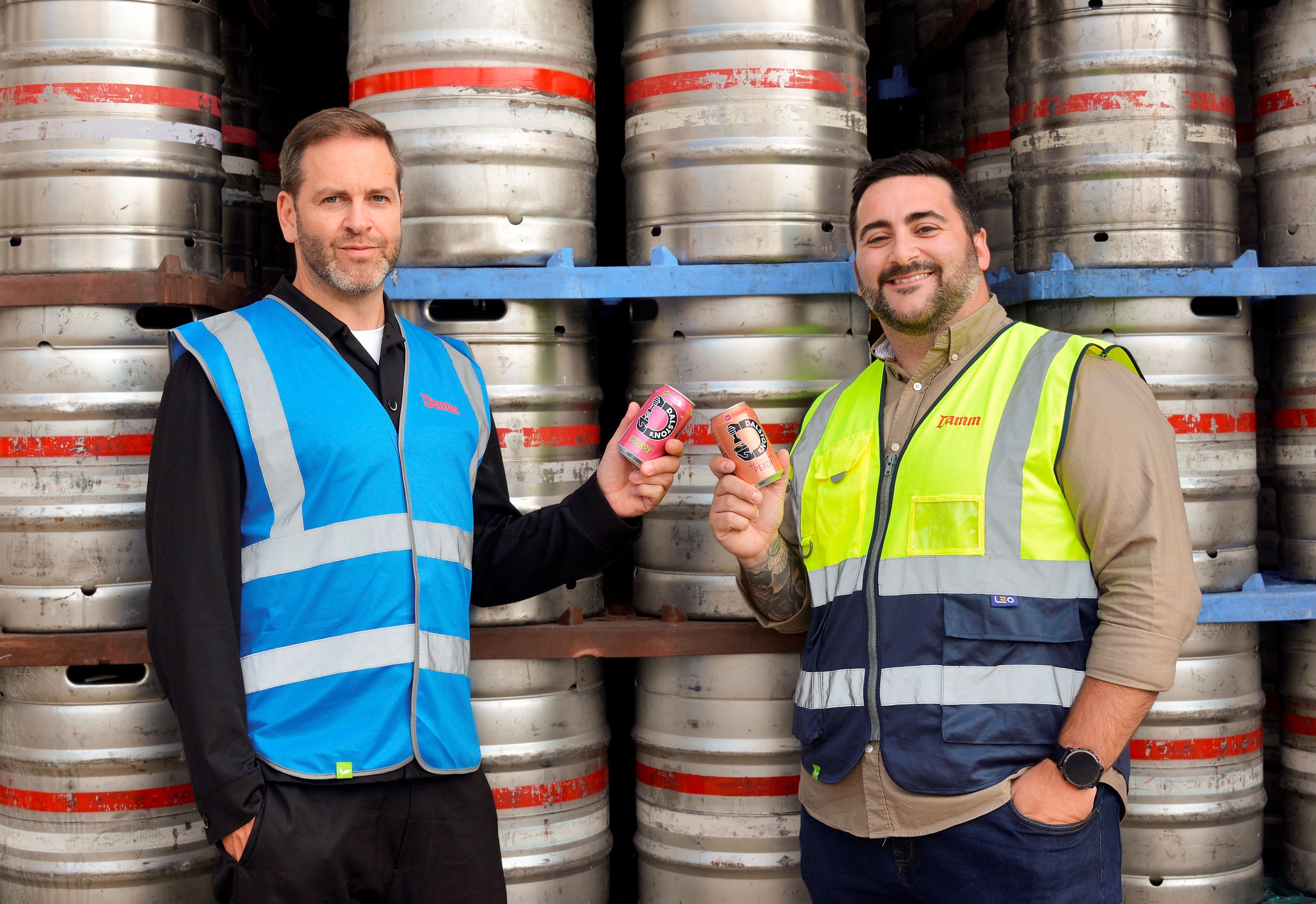 Two men clinking cans of soda together in front of barrels.
