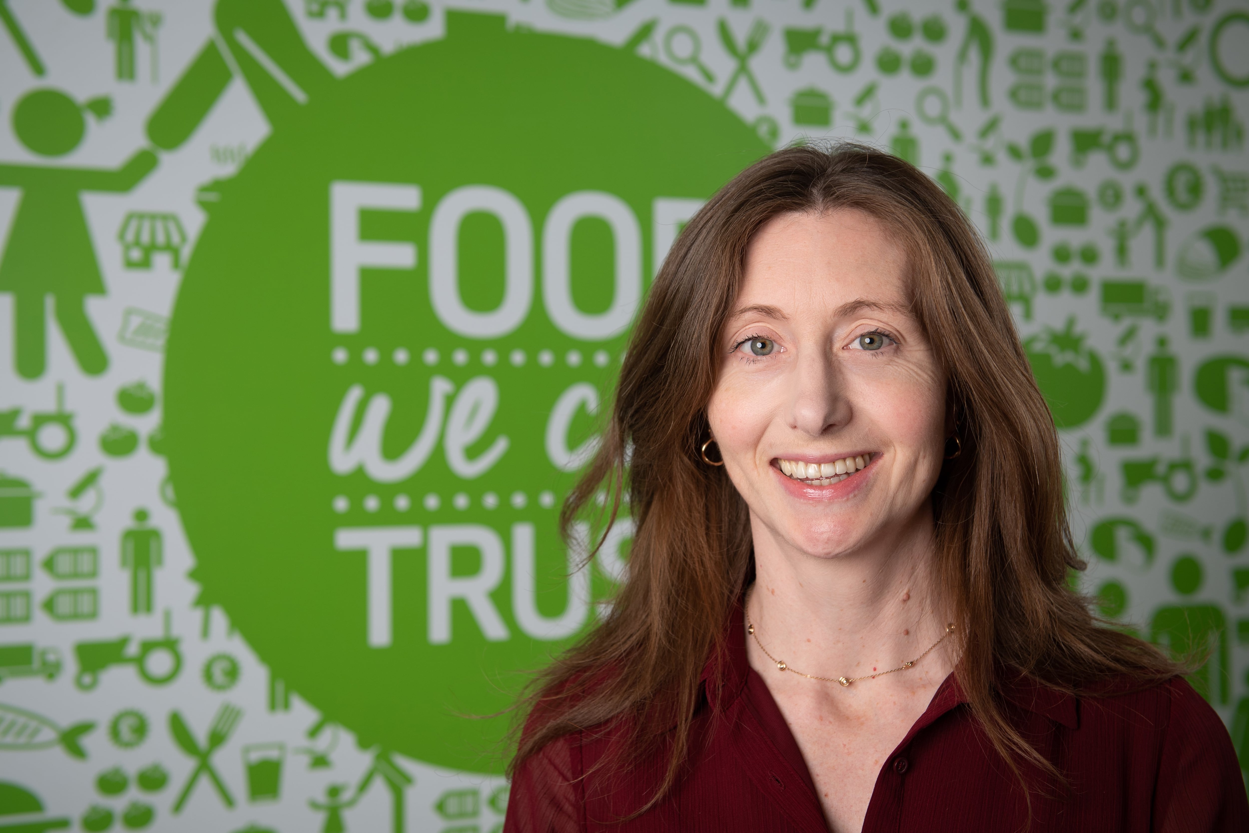 Middle-aged woman standing infront of a sign that reads 'food we can trust'. Woman is Katie Pettifer, FSA CEO